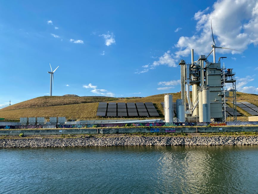 A landscape featuring a hillside with two white wind turbines and a series of black solar panels arranged in rows on the slope, set against a bright blue sky with scattered white clouds. In the foregr