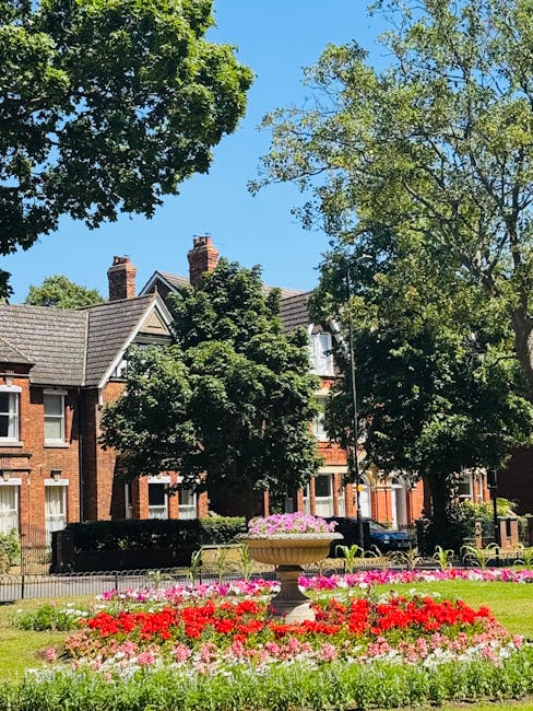 A well-maintained front garden in Mill Hill Village featuring a lush green lawn bordered by colorful flower beds with pink and red blooms, behind which is a decorative stone planter filled with pink f