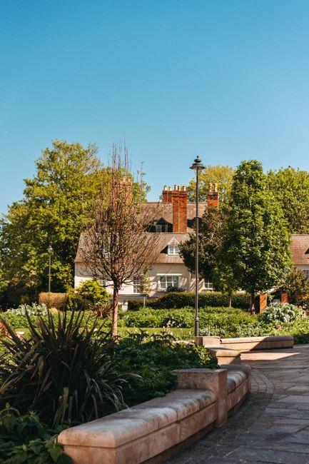 A well-maintained front garden in Mill Hill Village featuring a lush green lawn bordered by colorful flower beds with pink and red blooms, behind which is a decorative stone planter filled with pink f